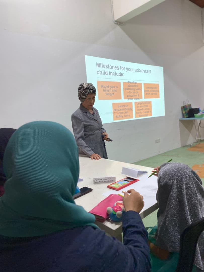 A teacher working with students at desks in a classroom, with a presentation on screen.